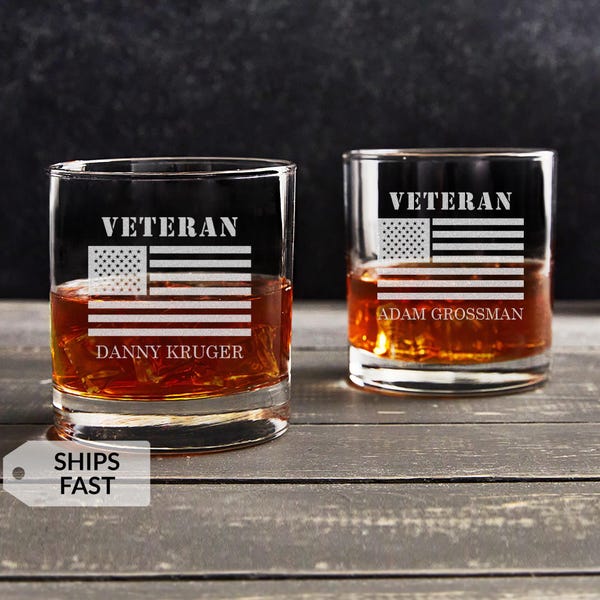 May include: Two clear whiskey glasses with etched designs. Each glass features the word "VETERAN" above a stylized American flag and a name below. The glasses are filled with amber liquid, likely whiskey, and sit on a wooden surface.