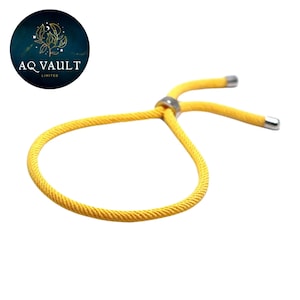 Pode incluir: Uma pulseira amarela ajustável com um nó deslizante e detalhes em metal prateado. A pulseira é feita de um material de cordão tecido. O logotipo da AQ Vault Limited está no canto superior esquerdo.