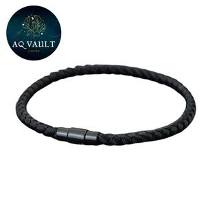 Op de afbeelding: Zwart gevlochten armband met een magnetische sluiting. De armband is cirkelvormig en lijkt gemaakt van een geweven materiaal. De sluiting is van donker, mat metaal. Het merklogo "AQ VAULT LIMITED" staat in de linkerbovenhoek.