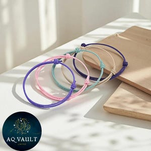 May include: A selection of cord bracelets in shades of purple, pink, teal, and beige. Each bracelet features a knotted fastening. The bracelets are arranged on a white surface, with a wooden board in the background. The logo "AQ VAULT LIMITED" is visible.