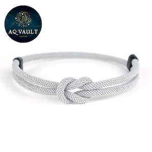 May include: A light grey rope bracelet with a knot design. The bracelet is made of a woven material and features black end pieces. The logo "AQ VAULT LIMITED" is in the upper left corner.