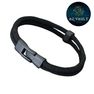 May include: A black bracelet with a woven rope design. The bracelet features a matte black clasp closure and a matching connector. The AQ Vault logo is visible in the upper right corner.
