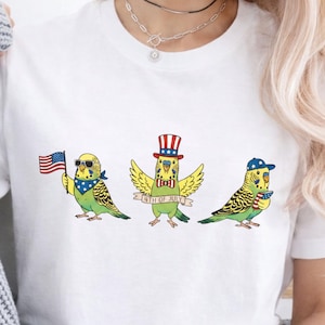 May include: White t-shirt featuring three cartoon parakeets celebrating the 4th of July. One parakeet holds an American flag, another wears a top hat, and the third wears a baseball cap. Each bird is adorned with patriotic accessories.