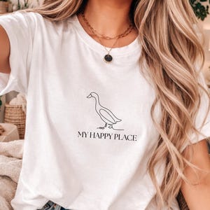 Puede incluir: Camiseta blanca con un dibujo lineal minimalista de un pato y el texto "MY HAPPY PLACE" debajo. El dise&ntilde;o es sencillo y elegante. Es una prenda informal para el d&iacute;a a d&iacute;a.