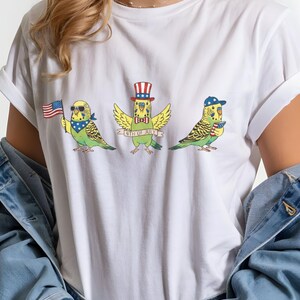 May include: White t-shirt featuring three cartoon parakeets celebrating the 4th of July. One parakeet holds an American flag, another wears a top hat, and the third wears a baseball cap. The text "4th of July" is on a banner.