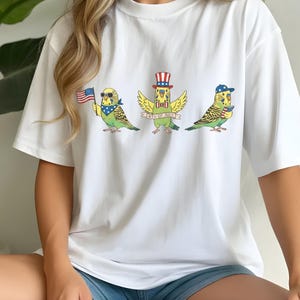 May include: White t-shirt featuring three cartoon parakeets celebrating the 4th of July. Each parakeet is adorned with patriotic accessories like sunglasses, a hat, and a bow tie, holding an American flag or a banner that reads "4th of July".