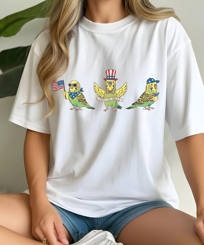 May include: White t-shirt featuring three cartoon parakeets celebrating the 4th of July. Each parakeet is adorned with patriotic accessories like sunglasses, a hat, and a bow tie, holding an American flag or a banner that reads "4th of July".