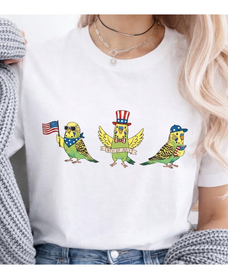 May include: White t-shirt featuring three cartoon parakeets celebrating the 4th of July. One parakeet holds an American flag, another wears a top hat, and the third wears a baseball cap. Each bird is adorned with patriotic accessories.
