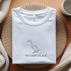 Puede incluir: Camiseta blanca con un dibujo minimalista de un pato y el texto "MY HAPPY PLACE" en negro. La camiseta est&aacute; doblada sobre una esterilla tejida. Fondo neutro, con una taza y una manta doblada.