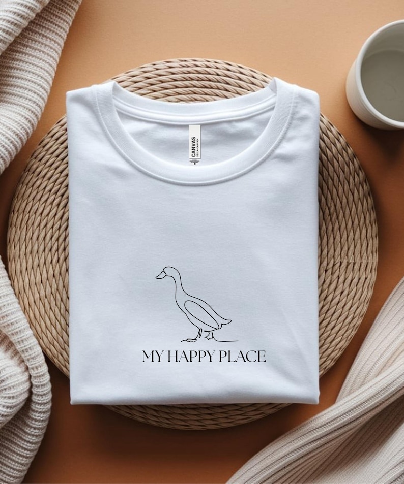 Puede incluir: Camiseta blanca con un dibujo minimalista de un pato y el texto "MY HAPPY PLACE" en negro. La camiseta est&aacute; doblada sobre una esterilla tejida. Fondo neutro, con una taza y una manta doblada.
