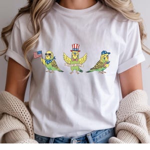 May include: White t-shirt featuring three cartoon parakeets celebrating the 4th of July. Each parakeet is adorned with patriotic accessories: sunglasses and a bandana, a top hat, and a baseball cap. The middle parakeet has a banner that reads "4th of July".