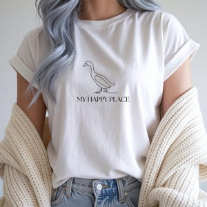 Puede incluir: Camiseta blanca con un dibujo lineal negro minimalista de un pato y el texto "MY HAPPY PLACE". La camiseta tiene las mangas dobladas y se combina con unos vaqueros azules. Un su&eacute;ter color crema est&aacute; drapeado sobre los hombros.