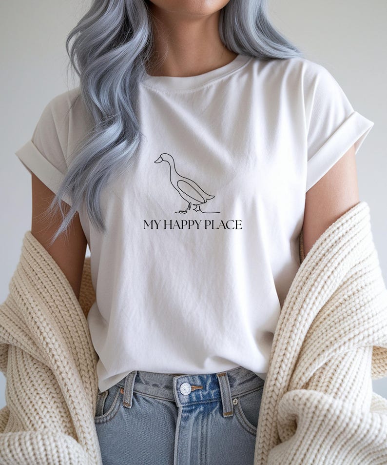 Puede incluir: Camiseta blanca con un dibujo lineal negro minimalista de un pato y el texto "MY HAPPY PLACE". La camiseta tiene las mangas dobladas y se combina con unos vaqueros azules. Un su&eacute;ter color crema est&aacute; drapeado sobre los hombros.