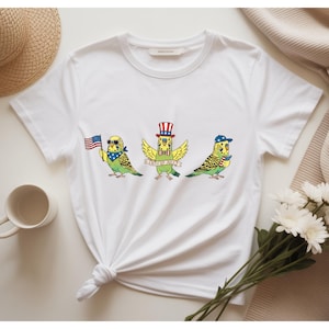 May include: White t-shirt featuring three cartoon parakeets celebrating the 4th of July. One bird holds an American flag, another wears a patriotic hat, and the third wears a baseball cap. The birds are green and yellow with blue accents.