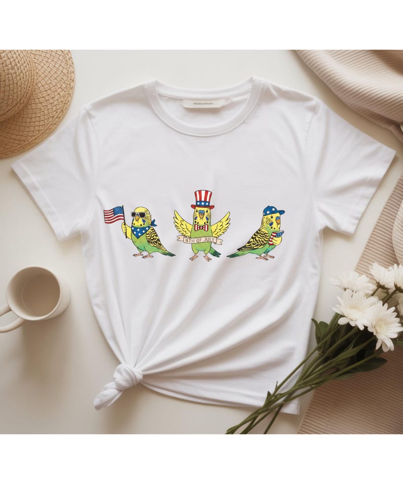May include: White t-shirt featuring three cartoon parakeets celebrating the 4th of July. One bird holds an American flag, another wears a patriotic hat, and the third wears a baseball cap. The birds are green and yellow with blue accents.