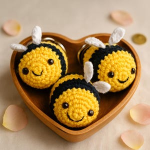 May include: Three handmade crochet bee keyrings in a wooden heart-shaped bowl. The bees are yellow and black with white wings and smiling faces. These keyrings are a charming, whimsical accessory.