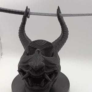 Könnte beinhalten: Eine mattschwarze Dämonenmasken-Skulptur mit zwei markanten, geschwungenen Hörnern und einem weit geöffneten Mund mit sichtbaren Fangzähnen. Die Maske ist auf einem runden schwarzen Sockel montiert und trägt ein langes, silberfarbenes Katana-Schwert.