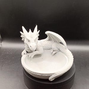 Könnte beinhalten: Hellgraue Drachenfigur mit schalenförmigem Sockel. Der Drache hat strukturierte Schuppen, Flügel und eine stachelige Krone. Die Figur steht auf einem schwarzen Drehständer vor dunklem Hintergrund.