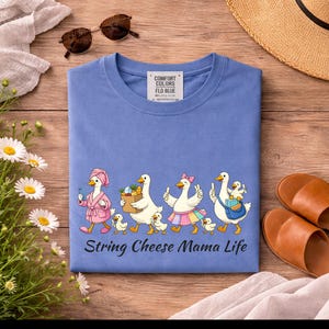 May include: A blue t-shirt with a whimsical illustration of geese, each with unique accessories, walking in a line. The text "String Cheese Mama Life" is below the geese. The shirt is surrounded by accessories.