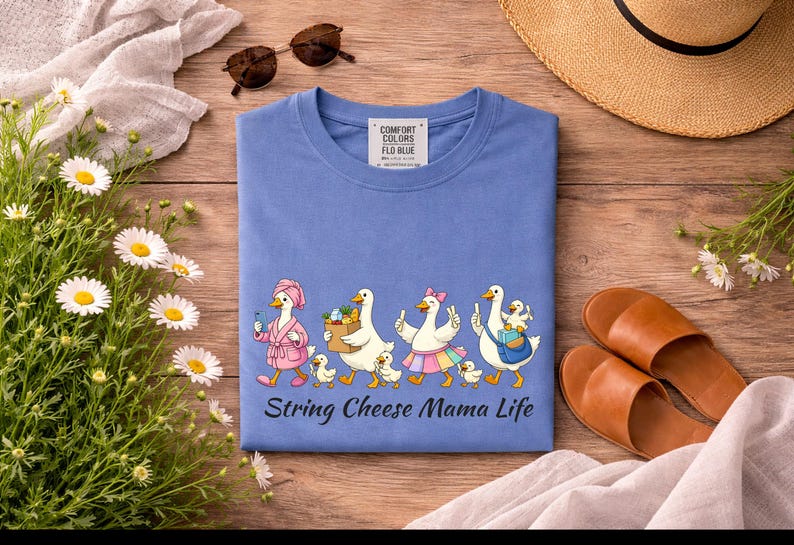 May include: A blue t-shirt with a whimsical illustration of geese, each with unique accessories, walking in a line. The text "String Cheese Mama Life" is below the geese. The shirt is surrounded by accessories.