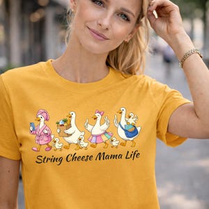 May include: Mustard yellow t-shirt featuring a whimsical graphic of geese in various outfits, each with ducklings. The text "String Cheese Mama Life" is below the graphic. The shirt is worn by a person.
