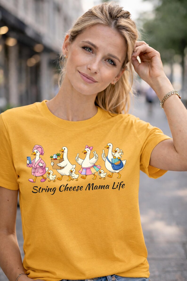 May include: Mustard yellow t-shirt featuring a whimsical graphic of geese in various outfits, each with ducklings. The text "String Cheese Mama Life" is below the graphic. The shirt is worn by a person.