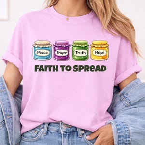 May include: Pale pink t-shirt with four illustrated jars labelled "Peace", "Prayer", "Truth", and "Hope". Below the jars, the text "FAITH TO SPREAD" is printed in dark green. The jars have colourful contents and lids.