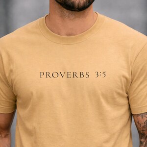 Op de afbeelding: Een beige t-shirt met ronde hals en de zwarte tekst PROVERBS 3:5 in een schreeflettertype in het midden van de borst. Het kledingstuk heeft korte mouwen en een standaard pasvorm, met een eenvoudig ontwerp en duidelijke, leesbare letters.