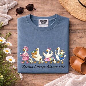 May include: A blue Comfort Colors t-shirt with a whimsical graphic of cartoon ducks, each with a chick, walking in a line. The text "String Cheese Mama Life" is below the graphic. The shirt is on a wooden surface with accessories.
