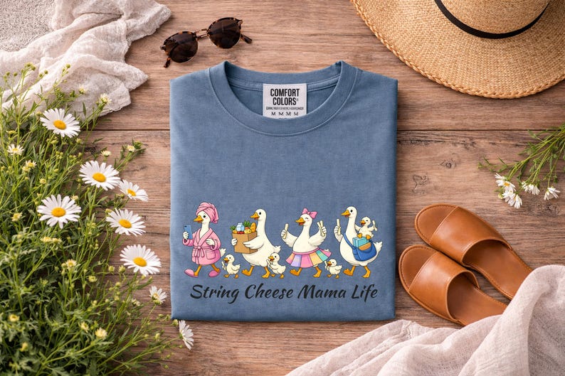 May include: A blue Comfort Colors t-shirt with a whimsical graphic of cartoon ducks, each with a chick, walking in a line. The text "String Cheese Mama Life" is below the graphic. The shirt is on a wooden surface with accessories.
