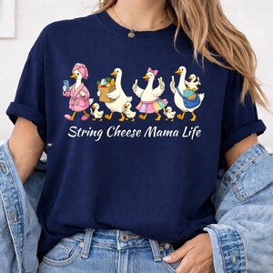 May include: Navy blue t-shirt featuring a whimsical illustration of geese in various outfits, including a bathrobe and a skirt. The geese are accompanied by ducklings. The text "String Cheese Mama Life" is printed below the illustration.