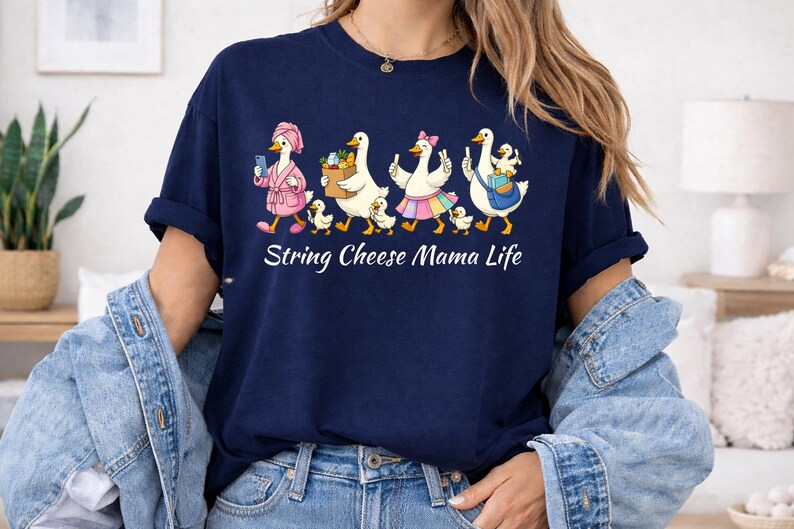 May include: Navy blue t-shirt featuring a whimsical illustration of geese in various outfits, including a bathrobe and a skirt. The geese are accompanied by ducklings. The text "String Cheese Mama Life" is printed below the illustration.