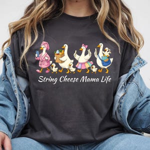 May include: Dark gray t-shirt featuring a whimsical illustration of geese. The geese are depicted in various outfits, including a pink robe and a skirt, with the text "String Cheese Mama Life" below the illustration.