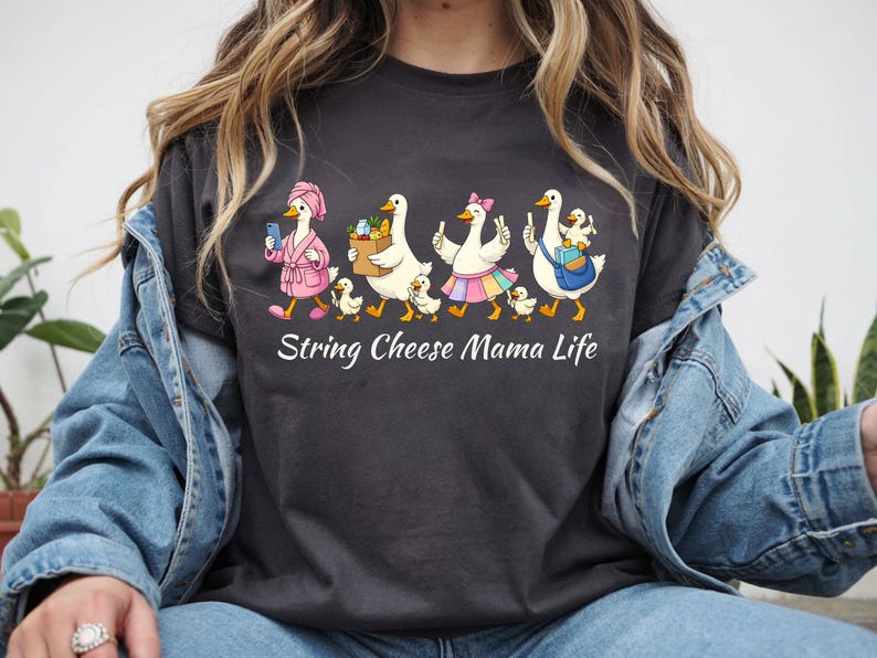 May include: Dark gray t-shirt featuring a whimsical illustration of geese. The geese are depicted in various outfits, including a pink robe and a skirt, with the text "String Cheese Mama Life" below the illustration.