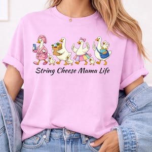 May include: A light pink t-shirt featuring a cartoon design of geese and goslings. The geese are dressed in various outfits and carrying items. The text "String Cheese Mama Life" is printed below the design.