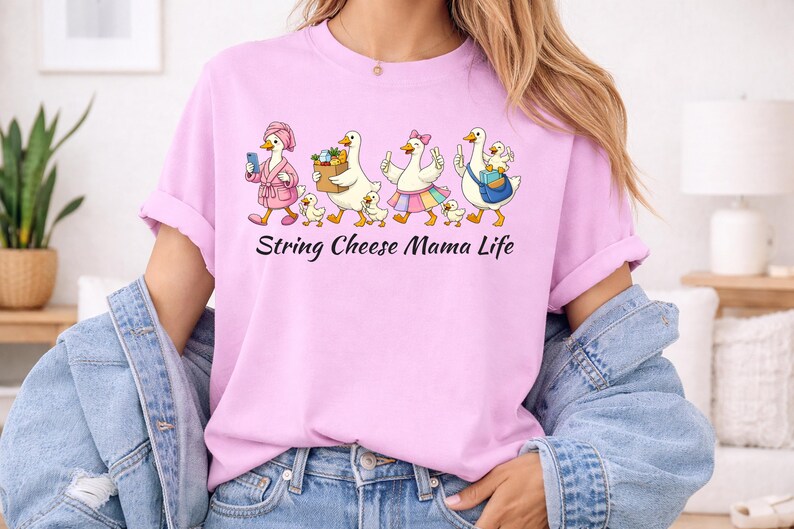 May include: A light pink t-shirt featuring a cartoon design of geese and goslings. The geese are dressed in various outfits and carrying items. The text "String Cheese Mama Life" is printed below the design.