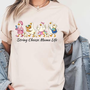 May include: Beige t-shirt with a whimsical graphic of geese and goslings. The geese are depicted in various outfits, carrying items like a phone, a basket of fruit, and a purse. The text "String Cheese Mama Life" is printed below the graphic.