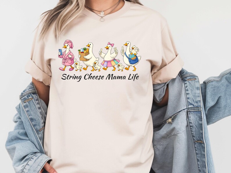 May include: Beige t-shirt with a whimsical graphic of geese and goslings. The geese are depicted in various outfits, carrying items like a phone, a basket of fruit, and a purse. The text "String Cheese Mama Life" is printed below the graphic.