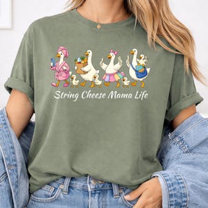 May include: Olive green t-shirt with a whimsical graphic of geese dressed in various outfits, walking with their goslings. The text "String Cheese Mama Life" is printed below the illustration. The shirt has short sleeves and a relaxed fit.