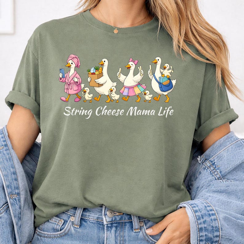 May include: Olive green t-shirt with a whimsical graphic of geese dressed in various outfits, walking with their goslings. The text "String Cheese Mama Life" is printed below the illustration. The shirt has short sleeves and a relaxed fit.