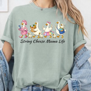 May include: A light green t-shirt with the text "String Cheese Mama Life". The shirt features a cartoon illustration of geese, each with unique accessories, walking in a line with their ducklings. The geese are wearing a pink robe, carrying groceries, and wearing a rainbow skirt.