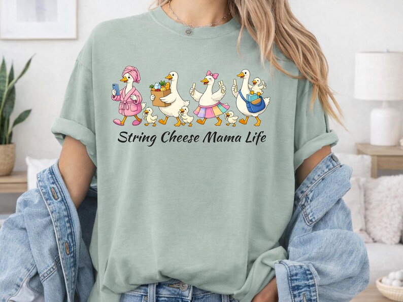 May include: A light green t-shirt with the text "String Cheese Mama Life". The shirt features a cartoon illustration of geese, each with unique accessories, walking in a line with their ducklings. The geese are wearing a pink robe, carrying groceries, and wearing a rainbow skirt.