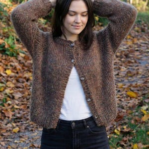 May include: A brown, button-down cardigan with a textured knit. The sweater features a round neckline and long sleeves. The buttons are light brown. The person is wearing a white top and black jeans.