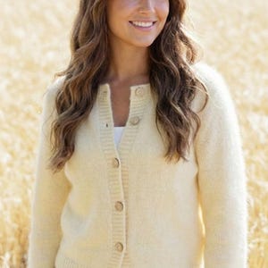 Può includere: Un cardigan giallo chiaro con chiusura a bottoni. Il maglione è realizzato in un materiale morbido e soffice e ha un design semplice. I bottoni sono marrone chiaro. La persona indossa un top bianco e jeans blu.