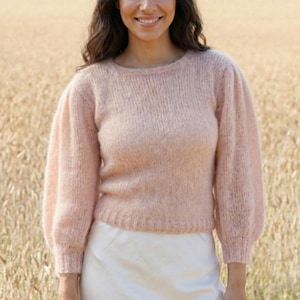 以下が含まれることがあります： 丸首の淡いピンク色のセーター。長くてパフスリーブが付いています。白いスカートと合わせています。女性は麦畑の前で微笑んでいます。