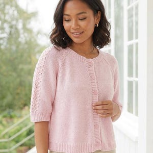 Könnte beinhalten: Hellrosa Strickjacke mit Knöpfen und kurzen Ärmeln. Der Strickpullover hat einen Rundhalsausschnitt und dekorative Details an den Ärmeln. Die Frau im Bild trägt die Strickjacke mit einer beigen Hose.