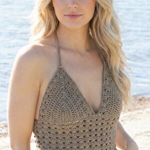 May include: A handmade, taupe-coloured crochet halter top with a deep V-neck. The top features a textured, open-weave design and a halter strap. The model is wearing white trousers. The background is a beach scene with the ocean.