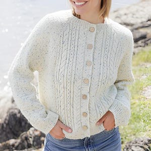 Peut inclure: Un gilet en tricot torsadé de couleur crème avec un motif moucheté composé de petites taches bleues et brunes. Le vêtement présente une encolure ronde, des manches longues à poignets côtelés et une fermeture boutonnée avec six boutons en bois.