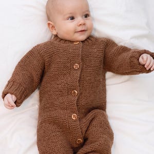 May include: A brown knitted baby romper featuring a textured garter stitch pattern, long sleeves with ribbed cuffs, and a front button placket with five small, round, light-colored wooden buttons. The garment has a simple crew neckline.