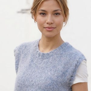 May include: A light blue knitted vest with a crew neck. The sleeveless top is made of a textured, soft material. A white t-shirt is visible underneath the vest. The model has blonde hair and is looking at the camera.